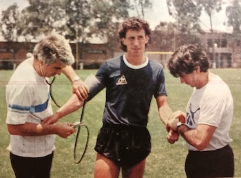 Tres hombres en un campo verde; uno con camiseta azul Le Coq Sportif es examinado por dos hombres, uno con un estetoscopio y otro palpando su muñeca