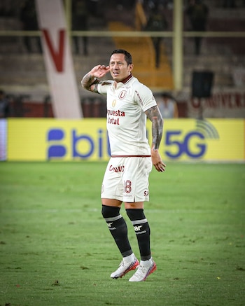 Un jugador de fútbol vestido con el uniforme blanco del club Universitario se lleva la mano a la oreja en la cancha