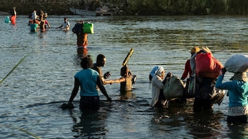 Imagen de un grupo de personas, incluidos adultos y niños, vadeando en un río con bultos sobre sus cabezas y hombros. Un bote se ve al fondo