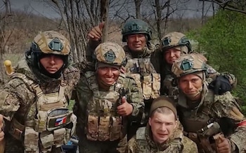 Seis hombres jóvenes con uniformes militares de camuflaje, chalecos tácticos y cascos posan juntos al aire libre, con vegetación y árboles al fondo