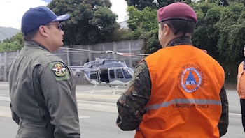 Dos hombres, uno con gorra azul y uniforme verde, otro con boina granate y chaleco naranja sobre uniforme camuflado. Un helicóptero gris en pista