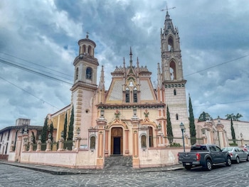 Fachada de iglesia con dos campanarios, techo ornamentado, paredes rosadas y beige, frente a una calle adoquinada con vehículos estacionados bajo cielo nublado