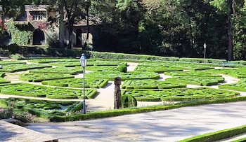Jardín con setos bajos que forman patrones, caminos, farola, estatua de madera y edificio cubierto de hiedra al fondo, con árboles