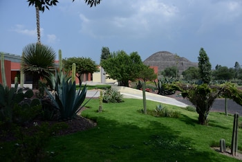 Jardín con césped verde, agaves y cactus, edificios modernos color terracota y amarillo, y una pirámide antigua de piedra al fondo bajo un cielo azul con nubes blancas
