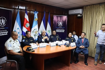 Siete hombres, varios con uniforme policial, sentados en una sala de conferencias alrededor de una mesa ovalada con banderas de Guatemala y un cartel oficial