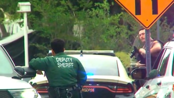 Dos oficiales de policía y varios coches patrulla con luces intermitentes en un escenario exterior; un oficial lleva un rifle, y el otro tiene "Deputy Sheriff" en su camisa
