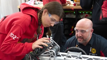 Joven con gafas de seguridad y sudadera roja examinando componentes de un robot. A su lado, un ingeniero calvo con barba y gafas observa
