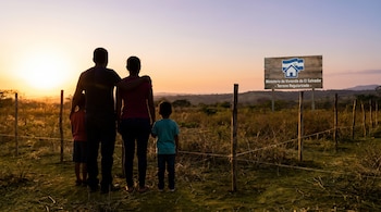 Siluetas de familia (dos adultos, dos niños) miran un campo cercado al atardecer. Un cartel del Ministerio de Vivienda de El Salvador indica "Terreno Regularizado".