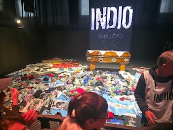 Ataúd de madera clara sobre un pedestal, frente a una pantalla grande con la palabra "INDIO" en letras blancas. Numerosas personas asisten, con objetos y flores coloridas en el suelo