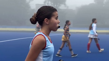 Una joven con uniforme de hockey azul claro y protector bucal oscuro mira hacia adelante en una cancha azul durante un día nublado, con otras jugadoras detrás