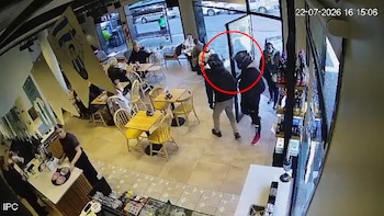 Interior de café con mesas, sillas y clientes. Dos hombres con casco entran, uno sujeta a un cliente. Empleado detrás de barra. Fecha y hora en la esquina superior derecha