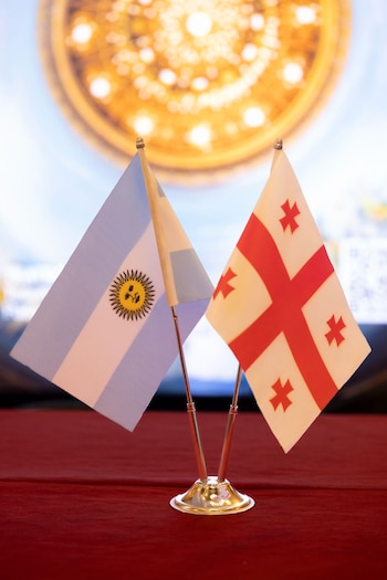 Dos banderas pequeñas, la de Argentina a la izquierda con el sol amarillo y la de Georgia a la derecha con cruz roja, en un soporte metálico sobre una tela roja