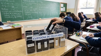 Una mano deposita un teléfono móvil dentro de una caja transparente etiquetada como custodia de celulares sobre un escritorio de aula.