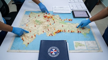 Un mapa de República Dominicana con marcadores de colores, manos con guantes azules que señalan zonas, una tableta y una carpeta oficial.