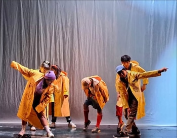 Un grupo de cinco bailarines, con gorros y abrigos impermeables amarillos, realizan poses dinámicas sobre un escenario oscuro con un telón blanco al fondo