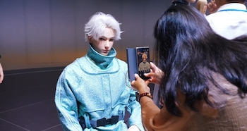 Robot humanoide con cabello blanco y ojos claros, vestido con un traje de color turquesa brillante. Una persona sostiene un teléfono móvil y lo fotografía