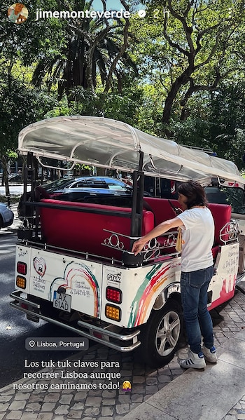Parte trasera de un tuk-tuk blanco con detalles rojos y coloridos, junto a una persona vestida de blanco, en una calle arbolada con adoquines