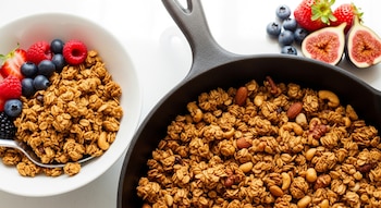 Granola dorada y crocante de avena y frutos secos en sartén y en bowl blanco, acompañada de fresas, arándanos, frambuesas e higos sobre fondo luminoso.