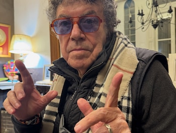 Piero luce gafas de marco naranja, bufanda a cuadros y abrigo negro mientras hace un gesto con ambas manos mirando a la cámara
