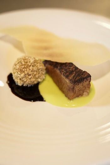 Corte de carne asado servido con salsa verde, salsa oscura, una esfera empanada y un crujiente sobre un plato blanco