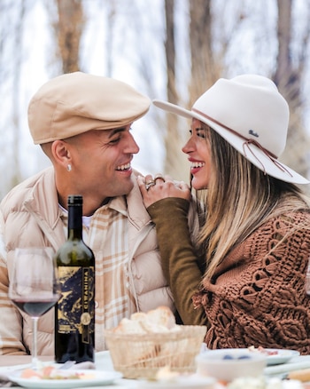 Un hombre y una mujer sonrientes se miran en una mesa al aire libre con una botella de vino tinto, copas, pan y platos de comida. El hombre lleva gorra y chaqueta, la mujer un sombrero