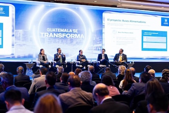 Cinco oradores en un escenario con una pantalla que muestra 'GUATEMALA SE TRANSFORMA' y 'Buses Alimentadores'; una audiencia se ubica de espaldas