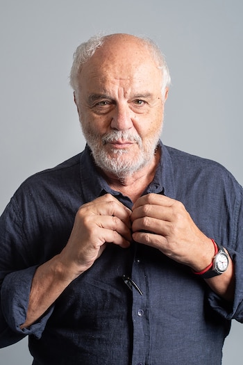 Retrato de Juan Leyrado, un hombre mayor de cabello y barba blancos, con una camisa azul oscura, abotonándose la camisa y mirando al frente