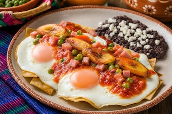 Plato con dos huevos fritos sobre tortillas, salsa de tomate, jamón en cubos, guisantes, plátano frito y frijoles negros cubiertos de queso.