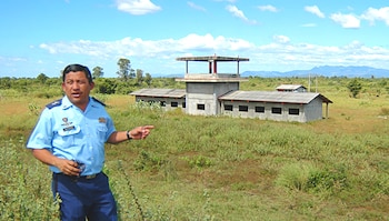 Un hombre con uniforme militar azul señala hacia una estructura de concreto en un terreno de vegetación baja con montañas al fondo