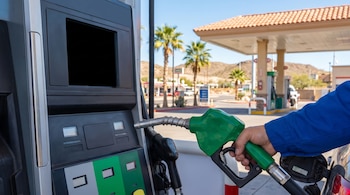 Mano de persona con camisa azul sosteniendo una boquilla de gasolina verde, llenando un coche en una estación de servicio. Se ven palmeras y montañas bajo un cielo claro.
