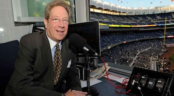 John Sterling con gafas y traje, sonríe frente a un micrófono en una cabina de transmisión; el Yankee Stadium, con sus asientos vacíos, se ve al fondo