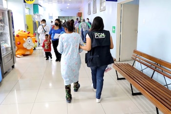 Pasillo de hospital con mujer de chaleco PGN, pacientes, niño, máquina expendedora, inflable naranja, banco de madera y señalización