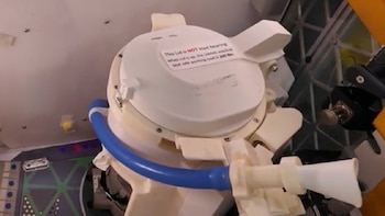 Primer plano de un inodoro espacial blanco con una tapa, un tubo azul en la base y una etiqueta de advertencia roja y negra en la parte superior