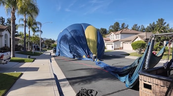 Vista de un globo aerostático azul y amarillo desinflado ocupando una calle residencial, con personas trabajando a su alrededor y casas con palmeras al fondo