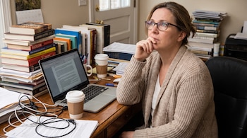 Mujer de mediana edad con gafas y suéter de punto sentada en escritorio de madera con un ordenador portátil abierto, pilas de libros, papeles y tazas de café.