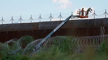 Dos trabajadores en una plataforma elevadora junto a una valla fronteriza alta con alambre de púas en la parte superior y en el suelo, con vegetación verde