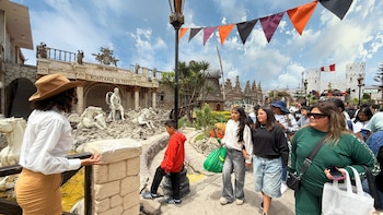 Grupo de personas camina en un complejo turístico con edificios temáticos, estatuas, una fuente, una bandera de Perú y adornos festivos bajo cielo nublado
