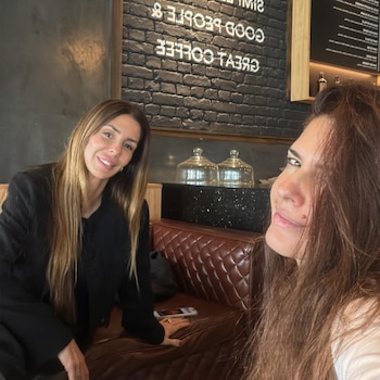 Dos mujeres sentadas en un sofá de cuero en una cafetería. Una sonríe a la cámara. La otra mujer de cabello castaño oscuro toma la foto. Pared de ladrillo con texto