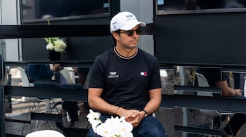 Un hombre con gorra blanca, gafas oscuras y camiseta negra está sentado frente a flores blancas con pantallas detrás.