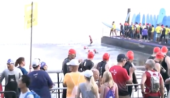 Nadadores con gorros rojos en el agua, personas en un muelle, espectadores en la orilla, una boya de demarcación y un cartel amarillo