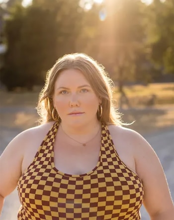 Retrato de Lindy West con cabello castaño claro, aros dorados, un collar con un pequeño dije, y un top a cuadros amarillo y granate