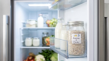 Interior de refrigerador moderno con puerta abierta. Frasco de vidrio Mason con avena y chía en estante de la puerta. Otros alimentos borrosos en el interior.