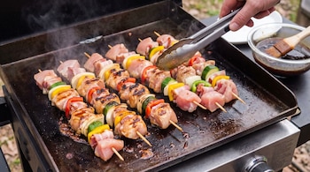 Vista aérea de brochettes de pollo, pimientos, cebolla, zucchini y panceta cocinándose en una plancha con humo. Una mano los gira con pinzas. Hay salsa teriyaki.