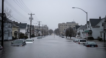 Vista de una calle de ciudad inundada con agua hasta la mitad de los coches aparcados. Casas residenciales a ambos lados y postes de servicios públicos.
