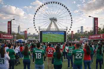 Multitud de aficionados con camisetas verdes y rojas ven un partido de fútbol en una pantalla gigante frente a la monumental rueda de la fortuna Aztlán en la CDMX.