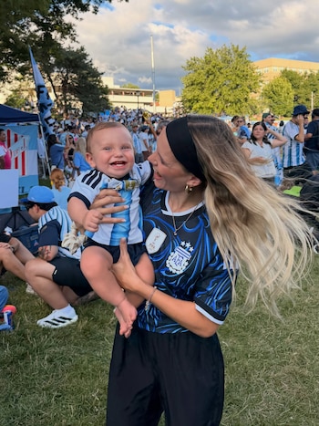 Una mujer rubia con camiseta de fútbol de Argentina sostiene a un bebé con camiseta del mismo equipo en un evento al aire libre