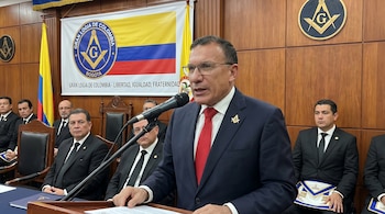 Roy Barreras en un podio con micrófono, viste traje oscuro, corbata roja y pin masónico. De fondo, bandera de Colombia y estandarte de la Gran Logia, con hombres en trajes.