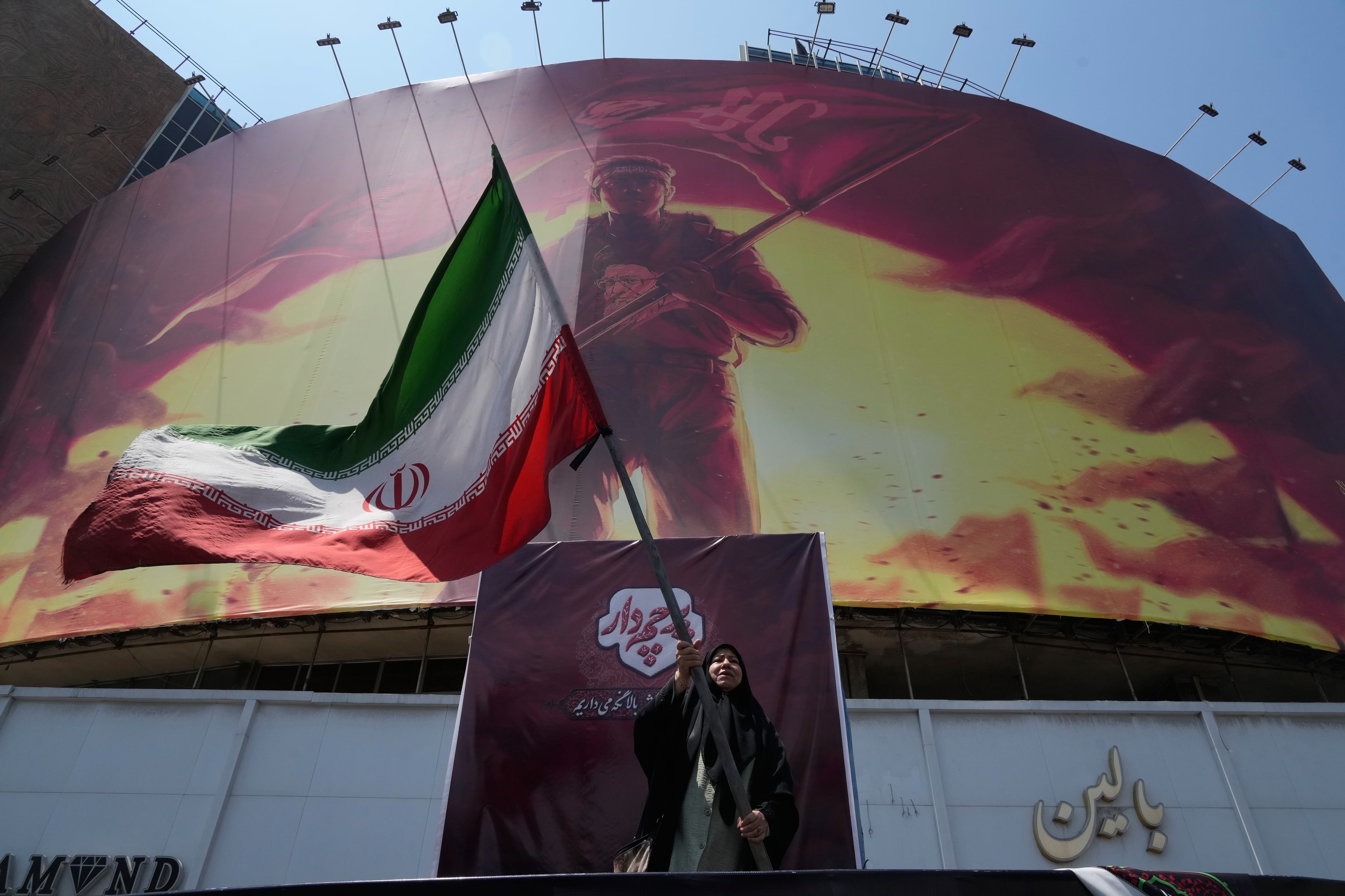Una mujer ondea la bandera iraní junto a una valla con la imagen del ayatolá Alí Khamenei, muerto el 28 de febrero, mientras Teherán condiciona la reapertura de Ormuz a que EEUU