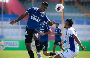 Un futbolista con uniforme azul y otro con uniforme blanco disputan un balón en el aire en una cancha de fútbol