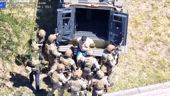 Vista aérea de vários oficiais em uniforme tático cercando um homem sem camisa em frente à janela do tanque.